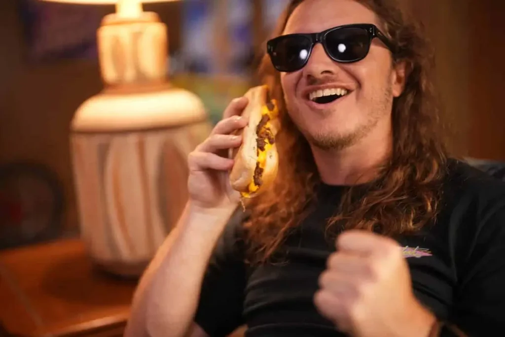 Man wearing sunglasses smiling while holding a cheesesteak sandwich to his ear like a phone