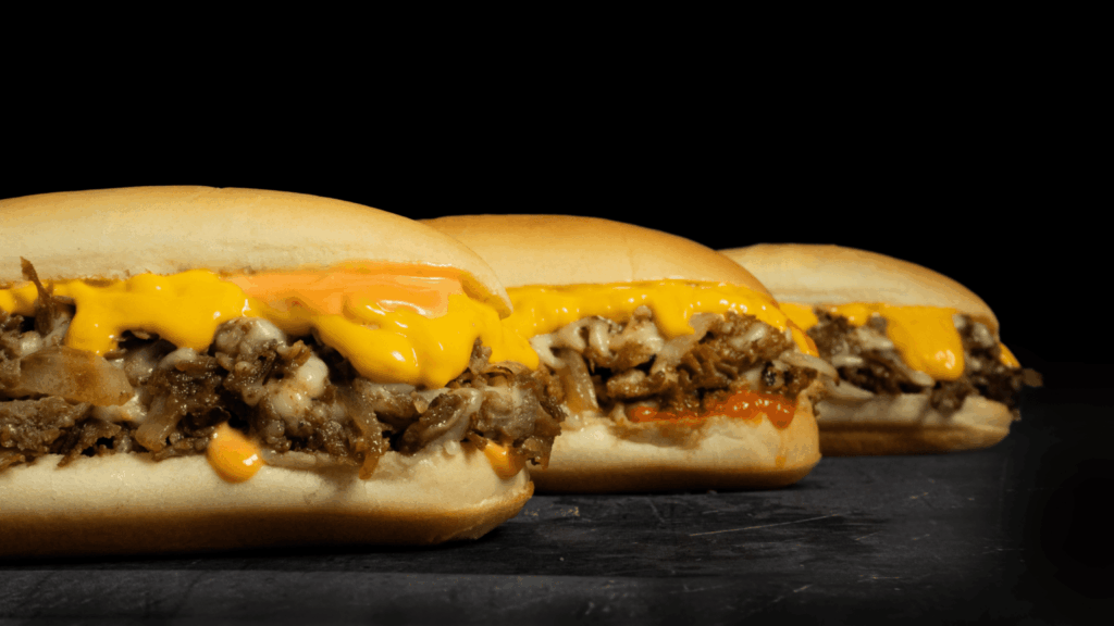 Three cheesesteak sandwiches with chopped beef, sautéed onions, and melted cheese on hoagie rolls against a dark background